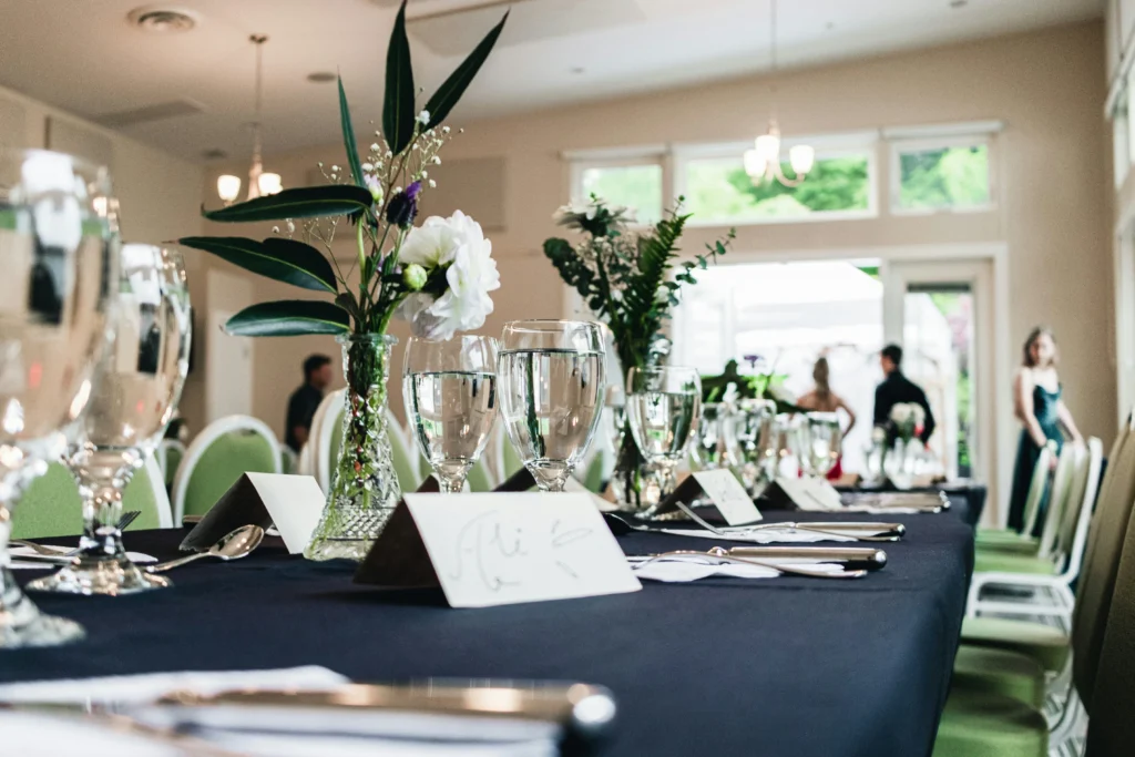 Eleganter Veranstaltungsraum mit liebevoll gestalteter Tafeldekorationen: weiße Blumengestecke, grüne Stühle, dunkelblaue Tischdecken und Champagnergläser in heller, moderner Eventlocation.